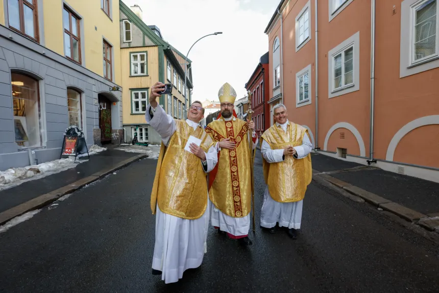Biskop Fredrik Hansen med mitra og bispestav i Akersveien, omgitt av to prester; den ene tar en selfie med biskopen