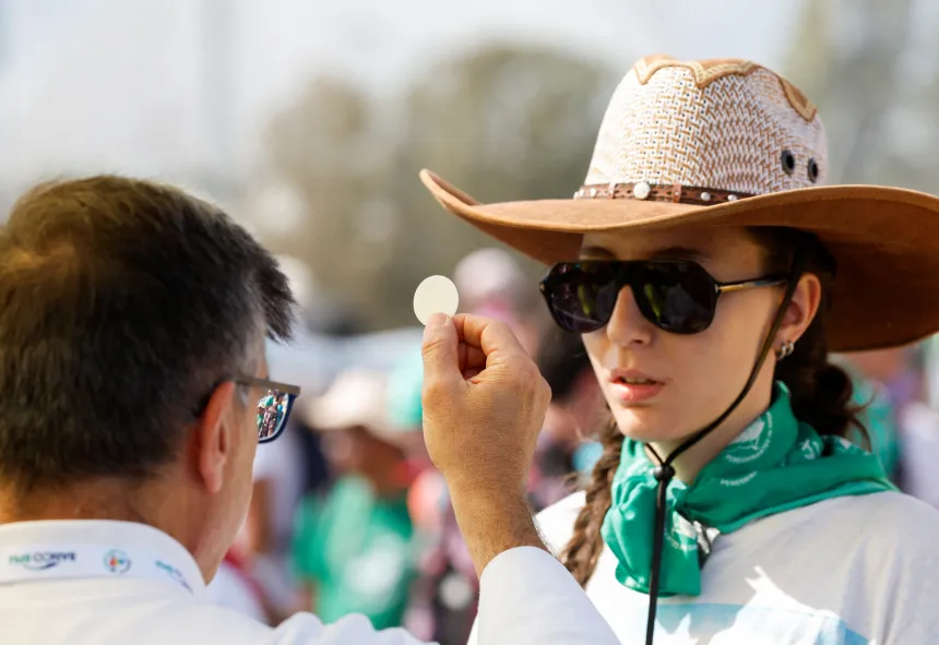 Ung kvinnelig pilegrim med hatt og solbriller mottar kommunion utendørs